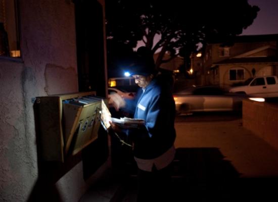 a male postal worker wearing a head lamp, looking at a stack of envelopes while delivering mail