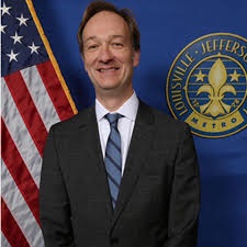 White man in a navy suit smiling while standing in front of an American flag and a seal for the city of Louisville, Kentucky