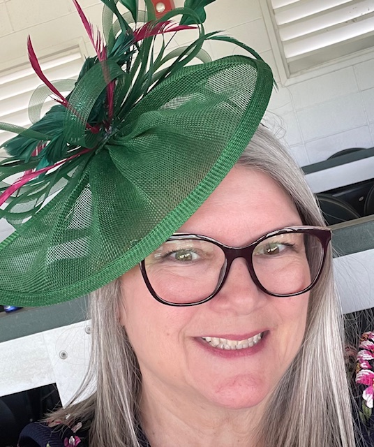 Image of a white woman with dark rimmed glasses in a green fascinator-style hat with pink feathers